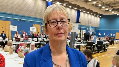 A woman with short blonde hair and thin black glasses wear a bright blue blazer while standing in a sports hall with people sat at a white table in the background.