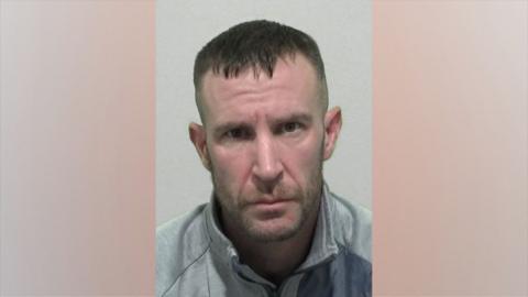 A mugshot of Kevin Belton. He has short, dark brown hair cropped at the sides and a stubbly but relatively neat beard and moustache. He is wearing a grey zipped jacket.