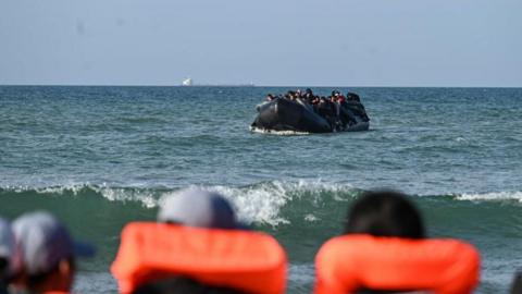 A stock image of migrants on a rubber dinghy. In the foreground people in caps and orange life jackets look out at them.