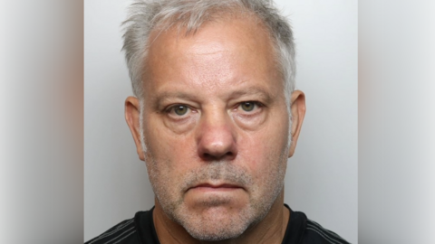 A mugshot of a man with short grey hair and wearing a black top.
