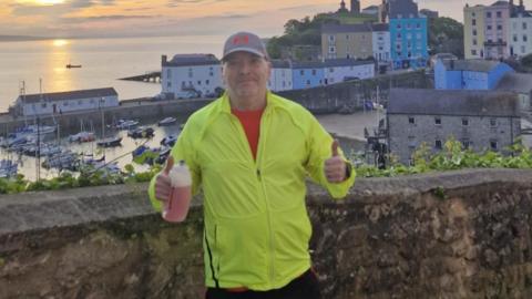 Darren Baker wearing a blue cap and yellow top putting his thumbs up at a harbour