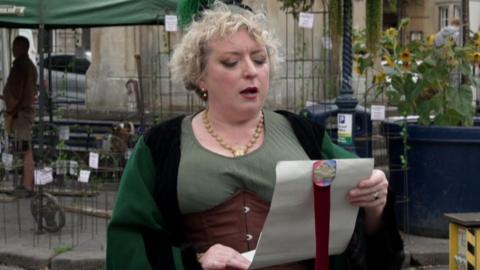 A woman with green robes shouts as she reads from a white scroll. She has short curly blonde hair and is standing in a town centre setting