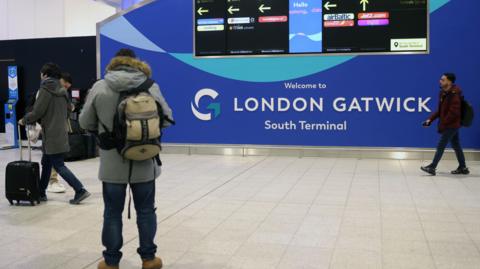 A man with a backpack on stands in front of a wall at Gatwick airport. The sign reads 'Welcome to London Gatwick South Terminal'. A screen shows directions to different airlines.  