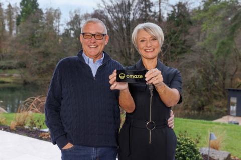 Nicola and her husband Martyn pictured outside their new home. Nicola holds a black card with the Omaze logo.