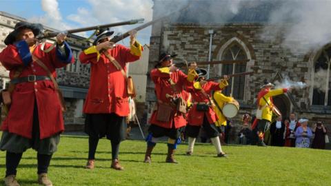 A line of people dressed in civil war-era uniforms firing muskets in front of Wimborne Minster church.