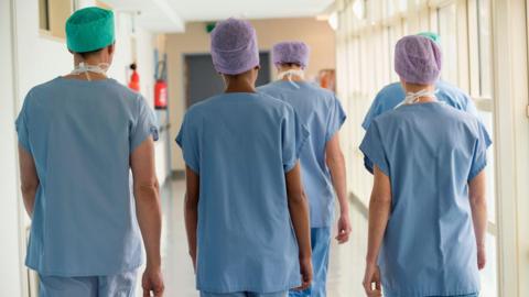 Five medical staff members walk through a hospital corridor. Their backs are to the camera. They are wearing blue medical scrubs and paper hats.