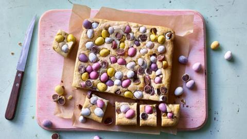 Uncut blondies topped with mini eggs on a pink board with a knife
