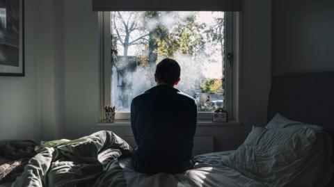 An anonymous man sitting on a bed looking out of the window