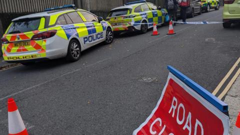 The turning into Cambridge Road has traffic cones across it and a sign reading 'road closed'. There are two police cars parked along it, an ambulance and an ambulance car.