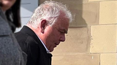 Allan Thursby leaving court. He has short white hair and is wearing a black coat.