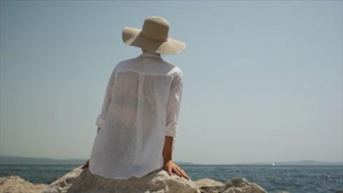 A woman sat in the sun wearing a loose shirt and sun hat