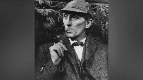 A black and white photo of a man in a cap and tweed jacket. He is smoking a pipe.