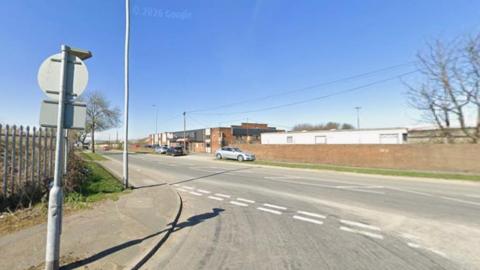 A wide road junction on a clear, sunny day. In the foreground, there is a pavement with a metal signpost and a tall security fence running alongside it. Several cars are driving along the road, which has white painted lane and junction markings.
On the right, a long brick wall lines the roadside, with low industrial buildings behind it.