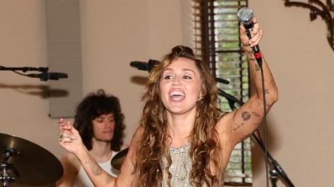Miley Cyrus on stage, holding a mic in the air. She has wavy long brown hair