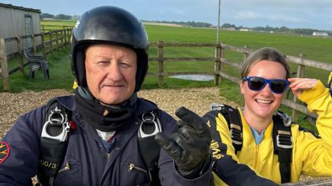 Chris Copeland wearing a black skydiving suit and black harness with metal carabiners. He is wearing a black helmet and holding up a rock symbol, while his granddaughter stands beside him leaning on the wooden fence. She is wearing a yellow skydiving suit and blue rimmed sunglasses.