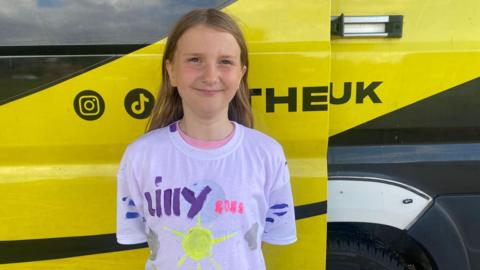 A young girl with brown hair is standing in front of a yellow van smiling at the camera, wearing a white t-shirt with paint on it which reads 'Lilly 2025'.