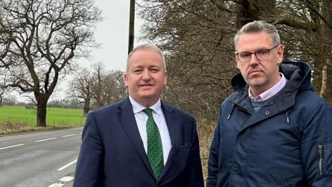 A man with short grey hair and a blue suit with a green tie and a man with grey hair and pink shirt in a blue coat standing beside a road with trees running alongside