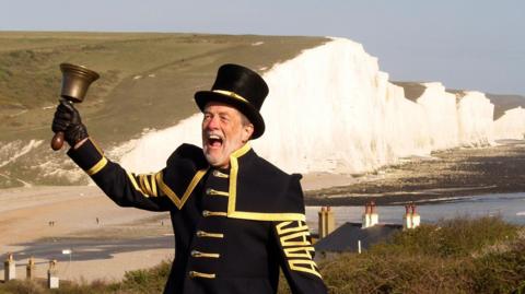 A man in a dark blue town crier uniform with gold trim and buttons, and top hat. He is holding a small bell on a handle and appears to be shouting. White cliffs and a beach can be seen in the background.