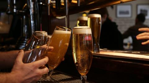 A bartender's hand holding glasses being filled with golden coloured larger from taps behind a bar with customers in the background. 