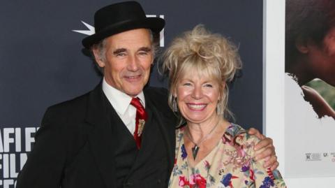 Claire van Kampen, who has a blonde updo and wears a floral dress with large silver earrings and a pendant necklace, smiles as her husband Mark Rylance puts his arm around her shoulder at a film screening 