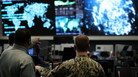 Two us cyber command officers in front of giant screens with data and information on them