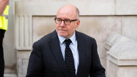 Attorney General Lord Richard Hermer leaves the Cabinet Office on Whitehall. He is wearing a dark suit and dark, spotted tie.