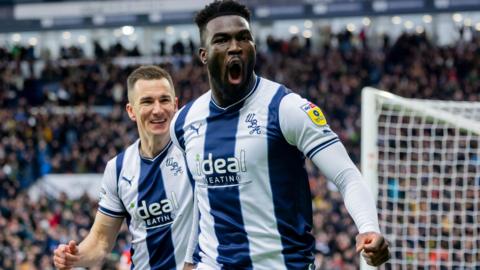Daryl Dike celebrates scoring for West Bromwich Albion. He is wearing a dark blue and white striped home shirt and is bellowing out as he starts his celebratory run. He is being chased by a smiling team-mate.