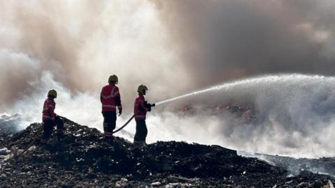 Three firefighters are wearing yellow helmets, red jackets and black trousers. They are standing on a pile of burnt rubbish on the left. One of them is holding a hose pipe trying to put out a fire.
