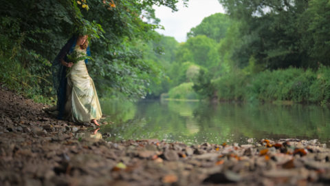 Megan Ruth-Trump is standing by the river. She is wearing a dress and is holding a bouquet of flowers. She is dipping her foot into the water.