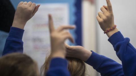 A generic photo of the back of several primary school children wearing blue jumpers. They have their hands up to answer questions.
