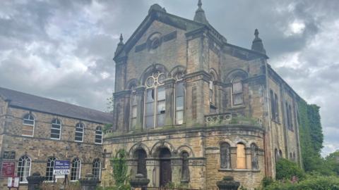 The picture is a large, historic church building constructed from yellowish-brown stone, featuring classical and Gothic revival architectural elements. The structure has a tall, rectangular form with a prominent central façade facing the street. The sky above is filled with dark, heavy clouds, giving the scene a slightly gloomy, dramatic atmosphere.
