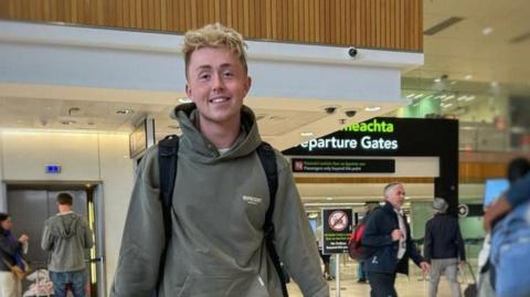 Jamie Harkin is standing in the airport departures area. He is wearing a green hoodie and wearing a black bag pack. There is an elevator behind him with two people standing outside of it. To his side there are four people with two hugging. In the background there is a large black departures gate.  