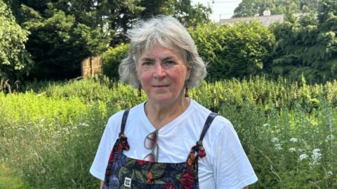 A woman with short grey hair is wearing a white t-shirt and dungarees, which have flowers on them. She is standing in front of wasit-high green plants and a few wildflowers. She has green framed sunglasses hanging from her t-shirt. 