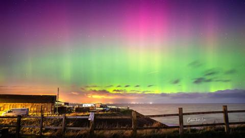 The Northern Lights over the sea at Mappleton in East Yorkshire with buildings and a fence in the foreground 