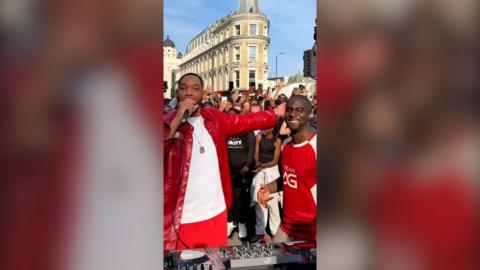 Will Smith holding a microphone besides another man, both wearing red, in front of a crowd.