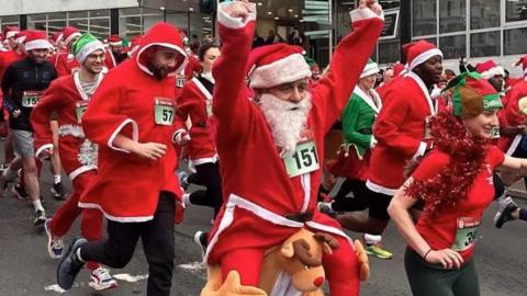 People dressed in red Santa jackets and hats. A man at the front has his arms aloft.