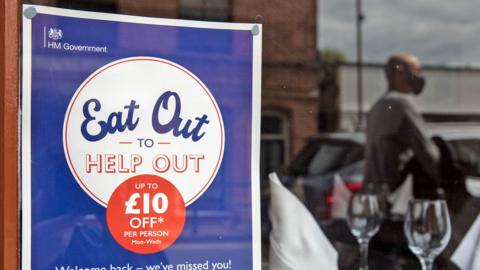 An Eat Out to Help Out poster in a window with two wine glasses and a blurred reflection of a customer