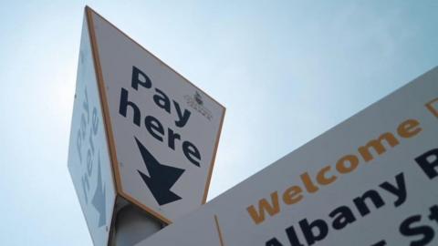 A close-up picture looking up at a Cornwall Council car park sign taken on a clear sunny day. The signs says 'Pay here' in bold black text. 