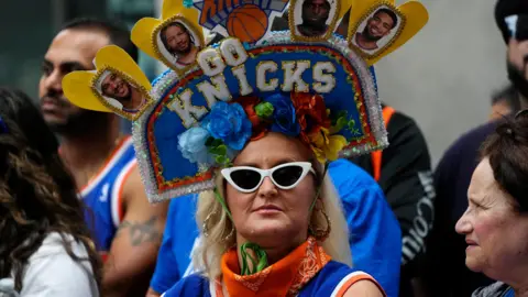 A woman dressed in blue and orange wears an extravagant Knicks hat and white sunglasses
