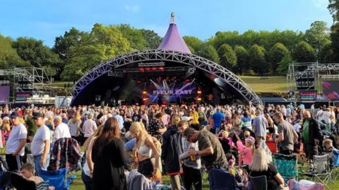 A large crowd gathered in front of a stage in Lower Castle Park. The stage is semi-circle shaped and has a purple roof. There are hundreds, if not thousands, of people stood in front of it.