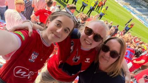 Three women taking a close-up photo at a football stadium. They are standing in the spectator seating area and are wearing red team jerseys and casual clothing; two are wearing sunglasses. Behind them, a green football pitch is visible with match stewards in high-visibility jackets and a large crowd of spectators, many also dressed in red. The scene appears bright and outdoors, suggesting a daytime sporting event.