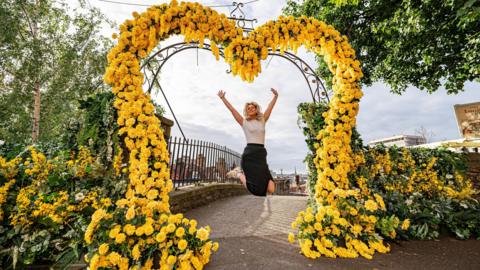 A woman jumping in the air with her arms up above her head and smiling. She is surrounded by a large yellow heart made of flowers