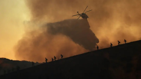 A helicopter drops water onto the fires in order to try and extinguish them as firefighters tackle the blaze on foot. They are surrounded by smoke. 