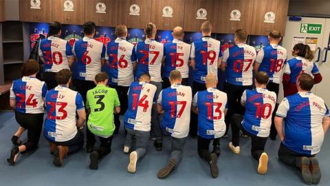 17 members of Dads FC kneeling in two rows in the changing rooms display the backs of their Rovers shirts - half blue, half white - with the names of their children on their shirts
