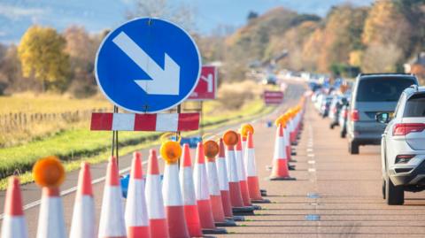 Orange traffic cones laid out to denote a lane is shut. There is congestion with rows of cars going down the road.