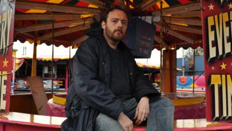 Dr Mitch Miller - a man with dark hair and a beard in jeans and a dark jacket sits on the edge of a stall at a fairground