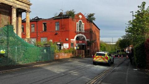 A police car is parked on a road next to a vacant pottery factory. The site is derelict and cones can be seen blocking the road in the distance.
