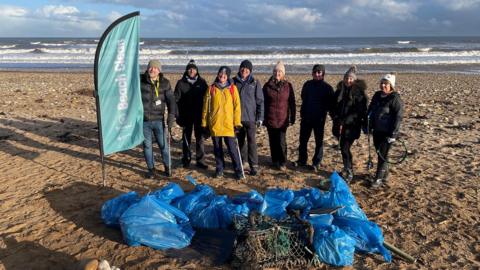 A group of eight people standing on a beach with blue bin liners full of rubbish - and the remains of a lobster pot -  in front of them. All are wearing warm-weather gear and some are carrying litter pickers. To their right is a turquoise banner labelled "Beach Clean". A rough sea and a cloudy sky are behind them.