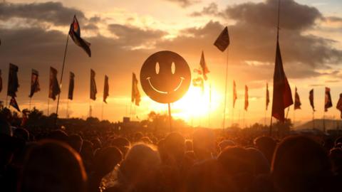 A smiley face held above a crowd at the Glastonbury festival with flags and a bright sunset in the background