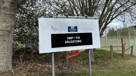 A sign outside a prison complex which reads "HM Prison Service: HMP/YOI Erlestoke". There are trees and grass behind it.